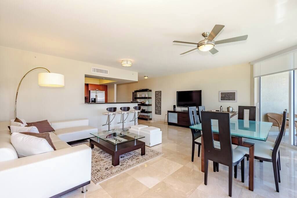 A modern living room with a glass coffee table, white sofas, a dining area with a glass table and chairs, a ceiling fan, and a kitchen in the background.