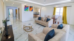 Una sala de estar moderna con un sofá color crema, una mesita de centro y un televisor sobre un mueble bajo.  Hay una zona de comedor con una mesa y sillas. La habitación tiene un ventanal con cortinas amarillas.