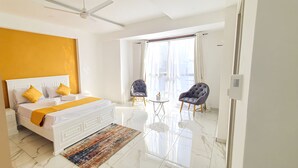 Una habitación de hotel moderna con una cama grande, un ventilador de techo, un balcón con dos sillas y un ventanal con cortinas translúcidas.