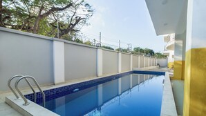 Una piscina con una escalera metálica, rodeada por un muro bajo y un edificio amarillo.