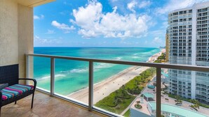 A balcony with a view of a beach and ocean, a wicker chair, and a high-rise building in the background. - Wow! Sunny Amazing Ocean Views - Great Location in Sunny Isles - Sleeps 5 (Sunny Isles Beach)