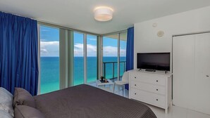 A hotel room with a large bed, a TV, a desk, and a view of the ocean.