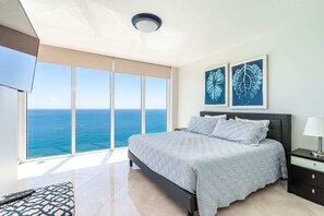 Quarto com cama, mesa de cabeceira, abajur e vista para o oceano através de amplas janelas.