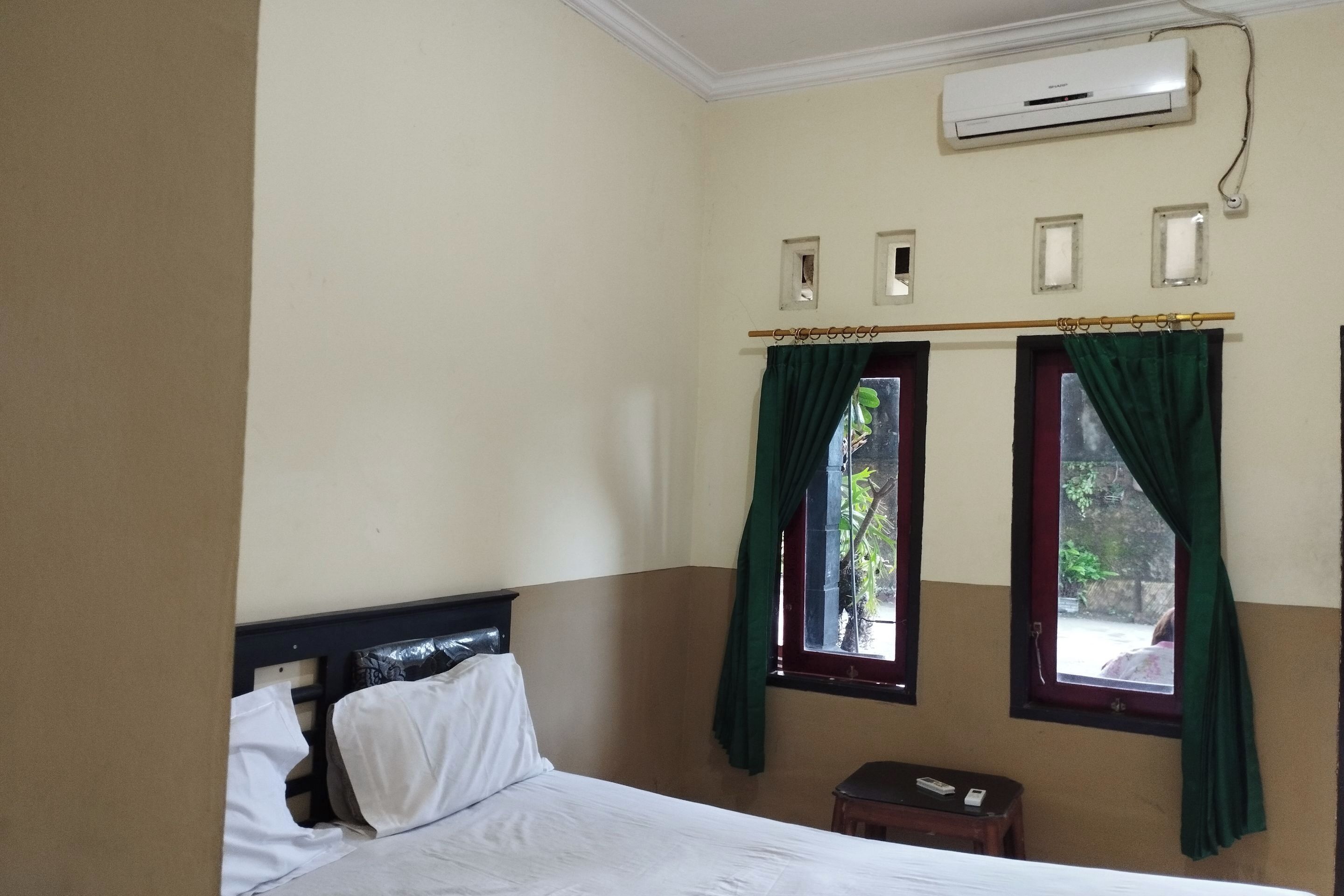 Een hotelkamer met een bed, nachtkastje, airconditioning en twee ramen voorzien van gordijnen.
