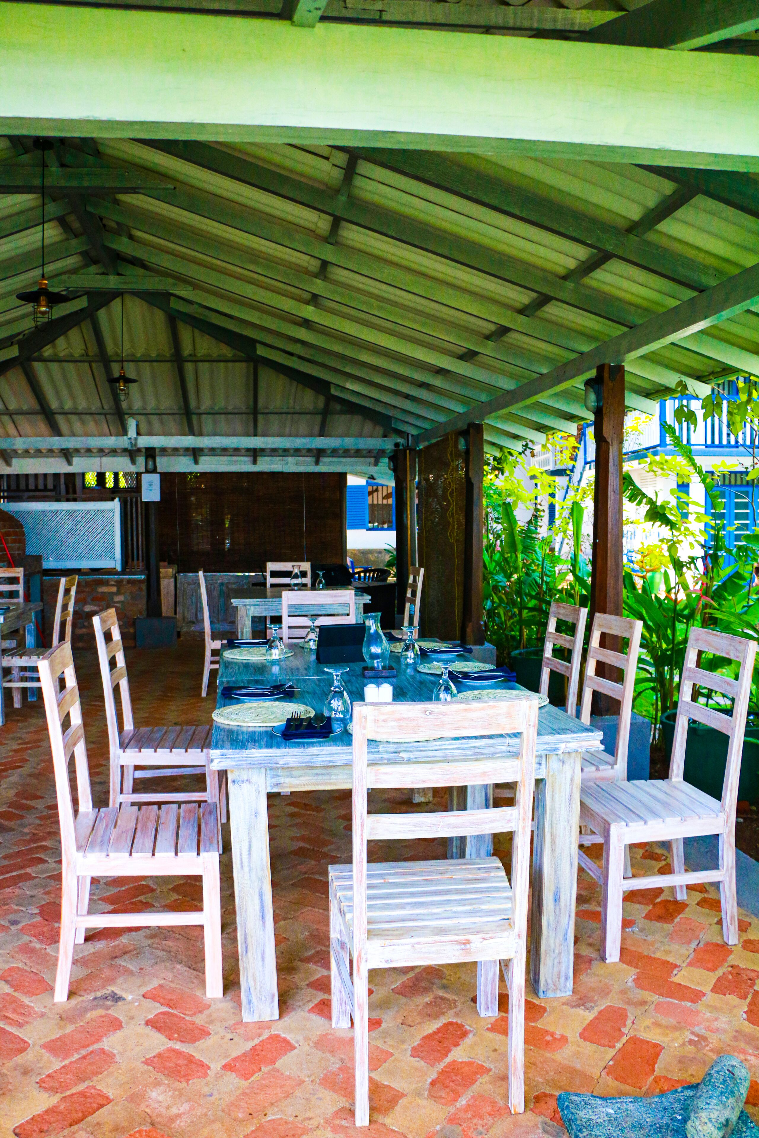 Un área de comedor al aire libre con techo cubierto, sillas y mesas de madera preparadas para comer.