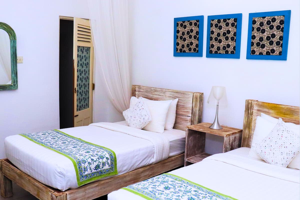 Twee houten bedden met witte lakens en groene accenten, een houten nachtkastje met een lamp, en een spiegel aan de muur.