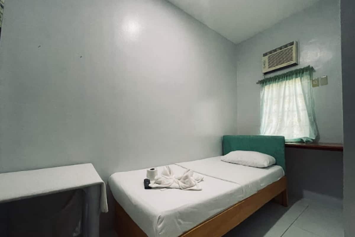 A single bed with a wooden frame and white bedding, a small table with a white tablecloth, and a window with a green curtain.