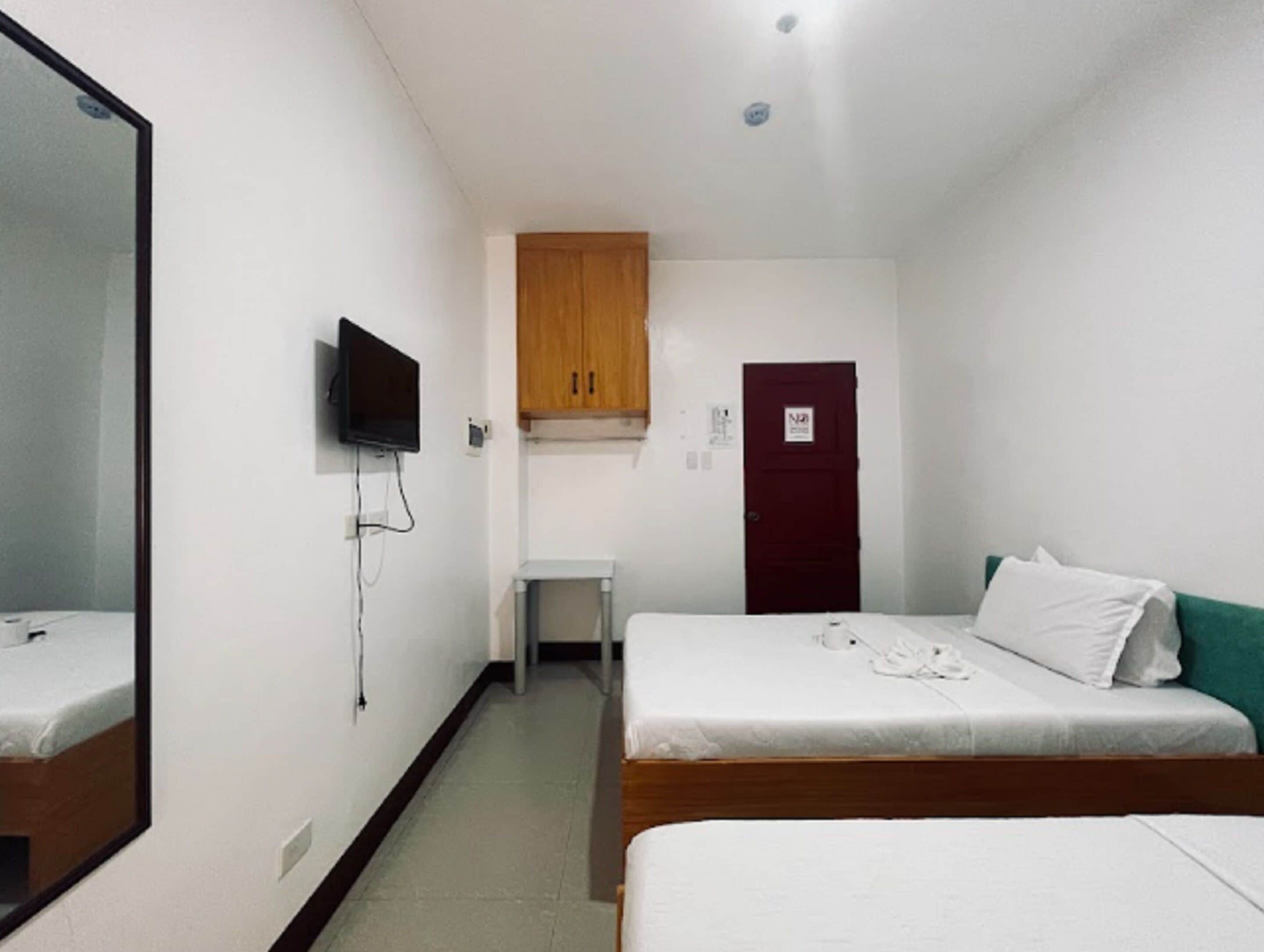 A hotel room with two single beds, a wooden wardrobe, a small desk, a television mounted on the wall, and a door with a sign.
