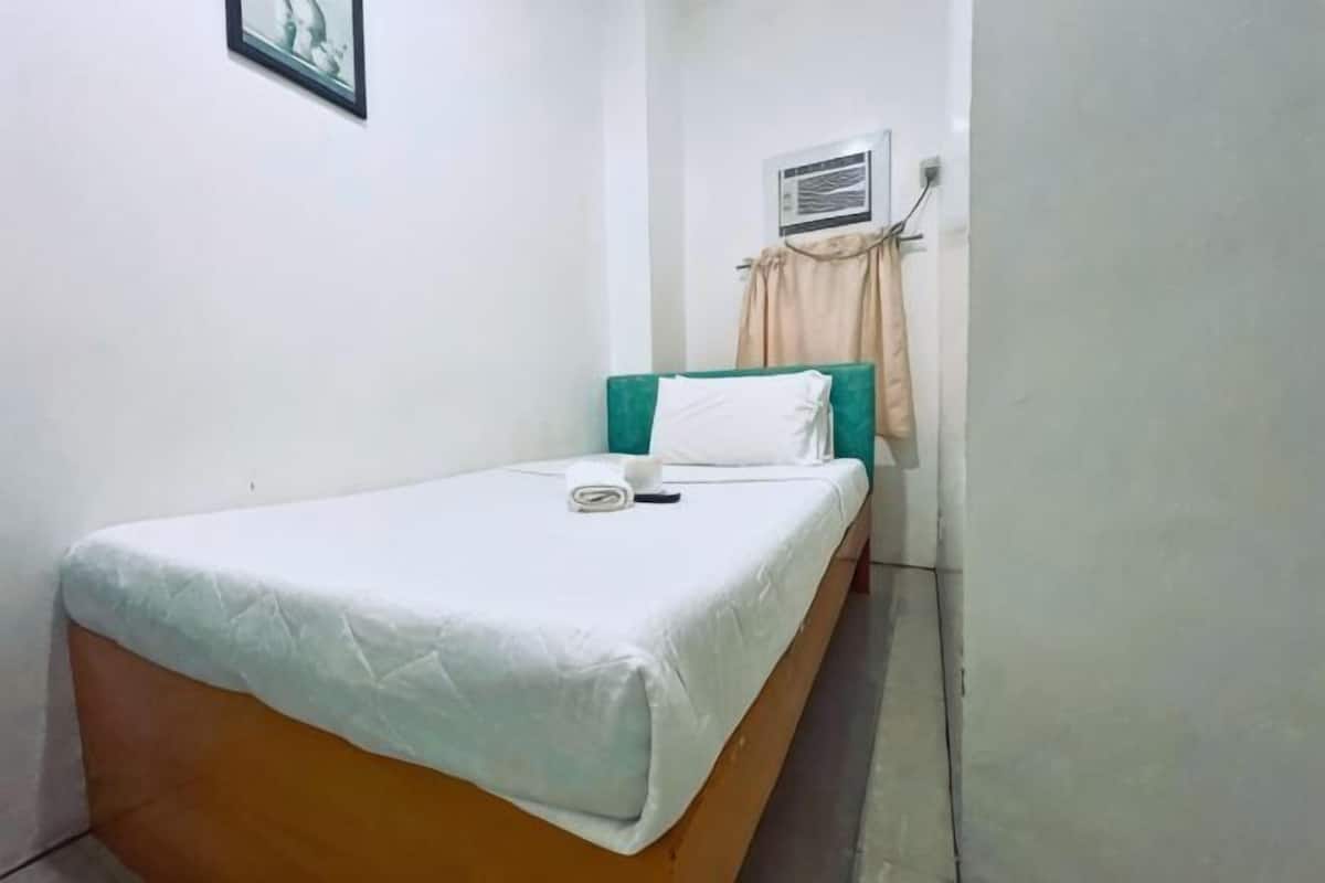 A single bed with a white mattress and teal headboard, a small bedside table, and a wall-mounted air conditioning unit.