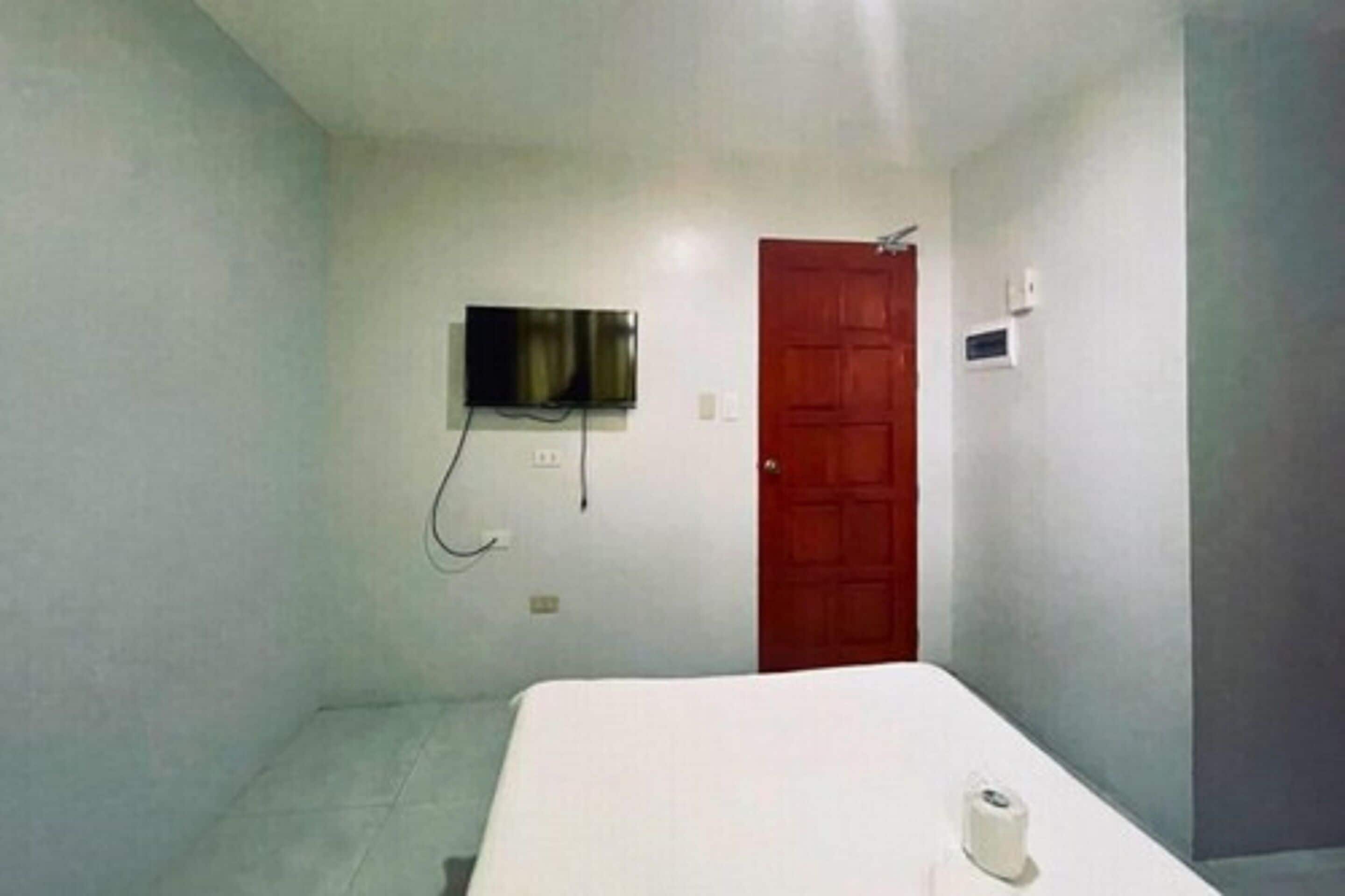 A small, simple room with a bed, a TV mounted on the wall, and a red door.