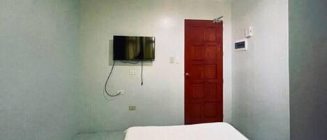 A small, simple room with a bed, a TV mounted on the wall, and a red door.