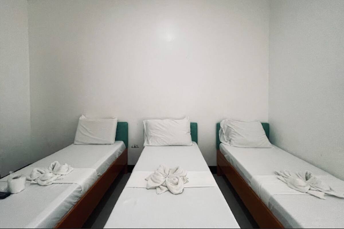 A room with three single beds aligned in a row, each with a white mattress and pillow, and a central bed with a towel arrangement.