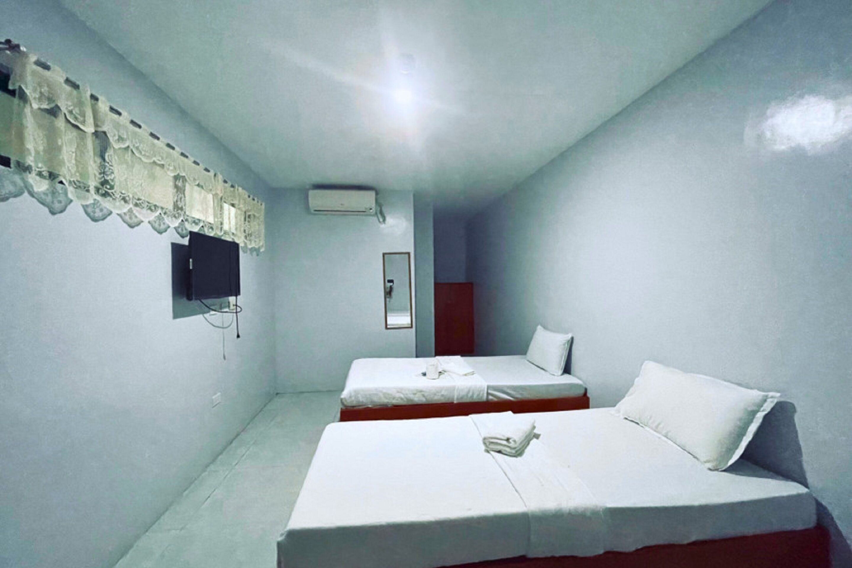 A small, simple room with two single beds, a TV mounted on the wall, and a window with a curtain.