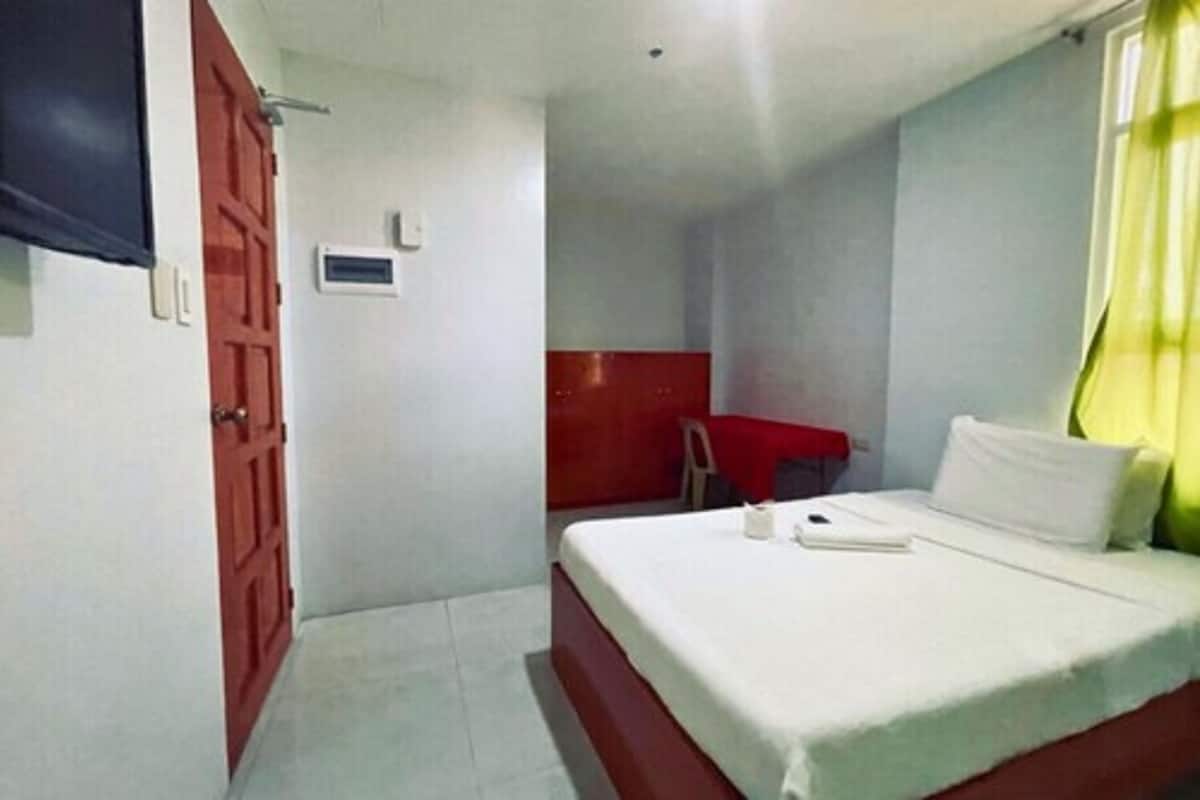 A small, single-bed room with a red door, a small desk, and a window with yellow curtains.