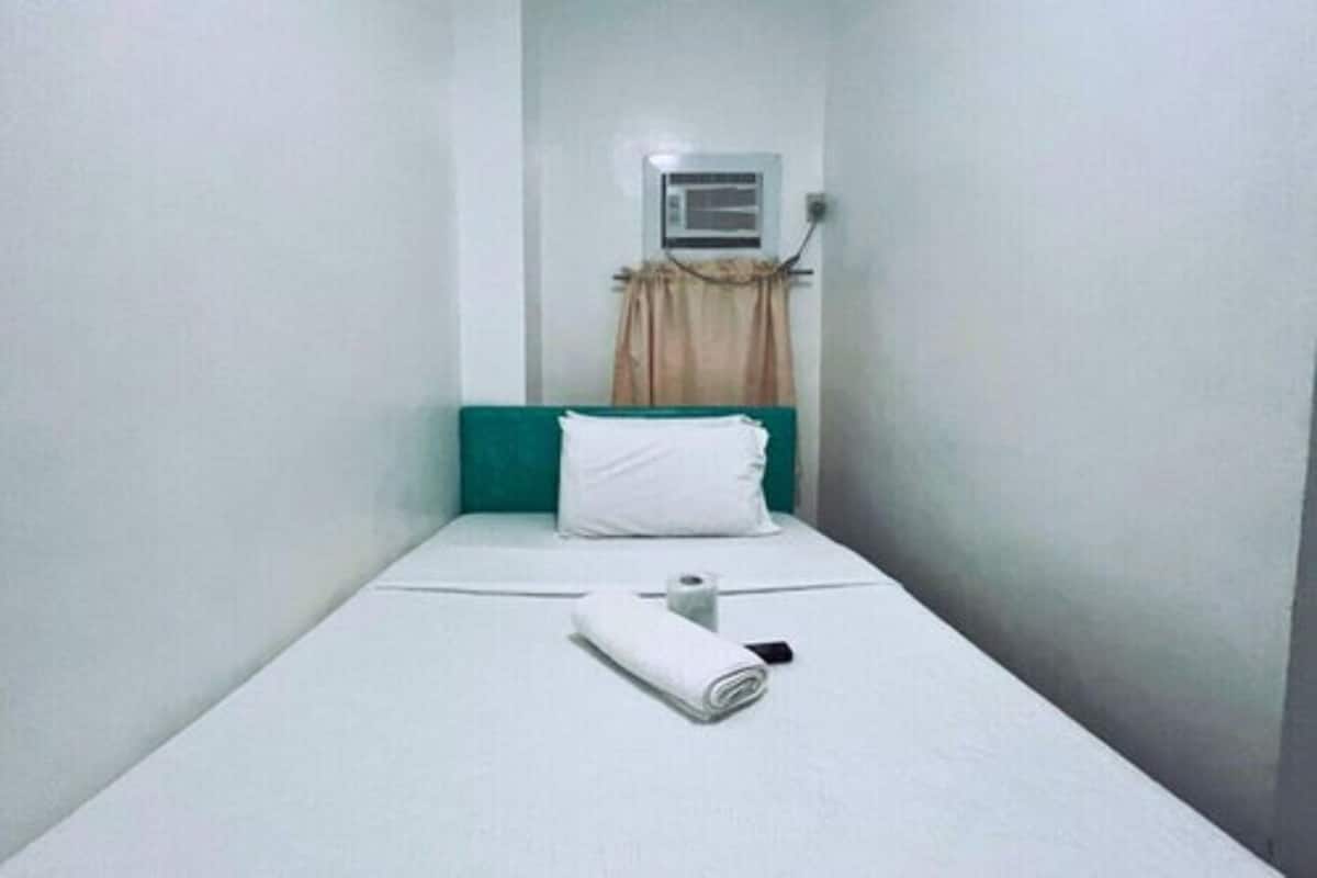 A small, narrow room with a single bed, a white pillow, a rolled-up towel, and a wall-mounted air conditioning unit.