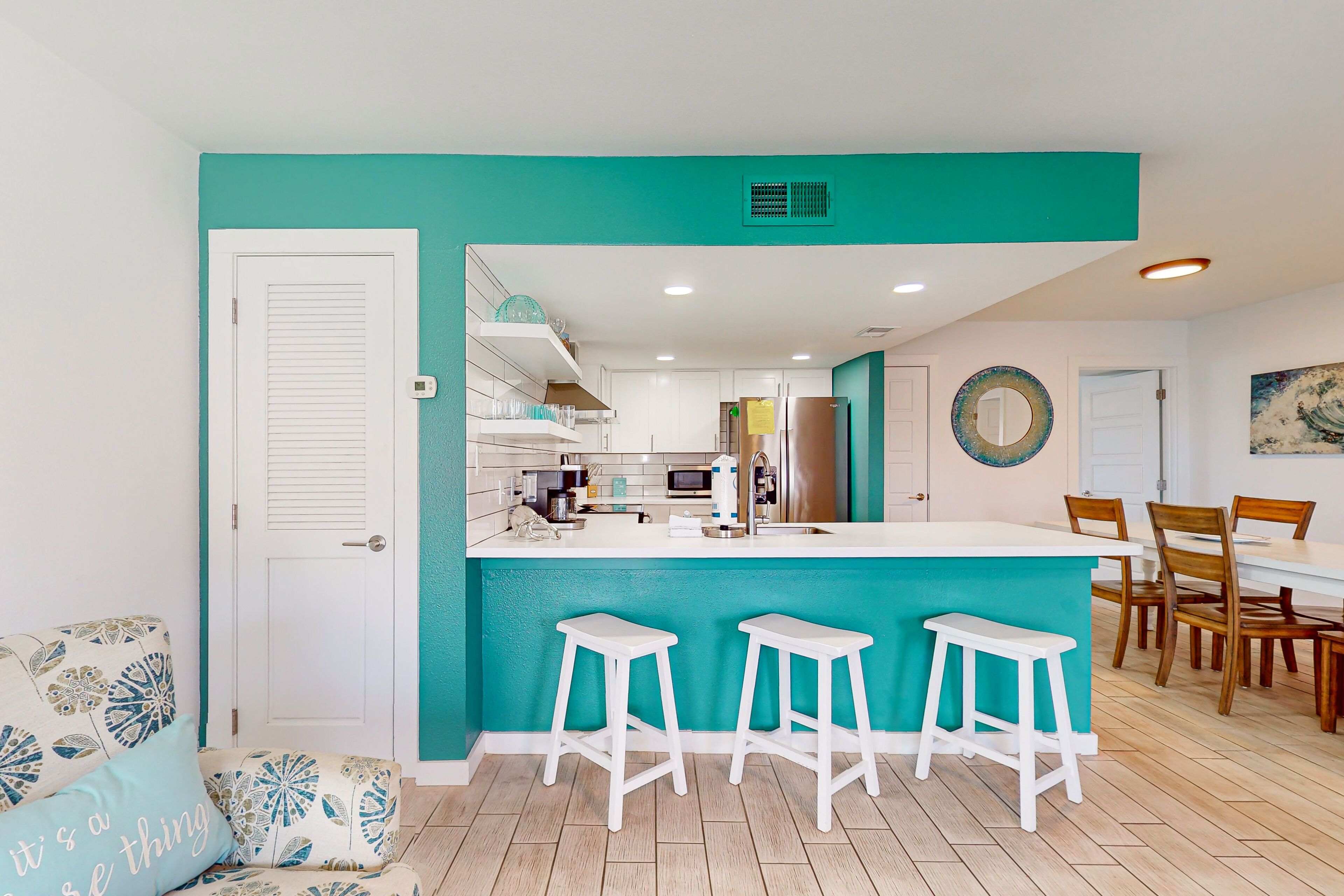 Une cuisine moderne avec un mur d’accent de couleur turquoise, des tabourets de bar blancs et un coin repas avec des chaises en bois.
