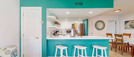 A modern kitchen with a teal accent wall, white bar stools, and a dining area with wooden chairs.
