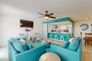 Un salon avec des canapés de couleur turquoise, un ventilateur de plafond et un coin cuisine équipé de tabourets et d’un bar.