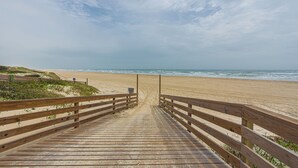 En træpromenade fører ned til en sandstrand, med havet i baggrunden.