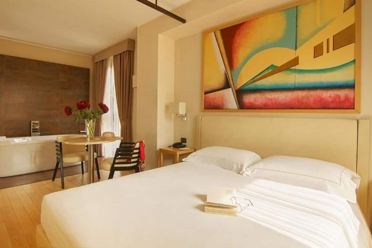 A hotel room with a large bed, a sitting area with a table and chairs, a bathtub, and a colorful abstract painting on the wall.