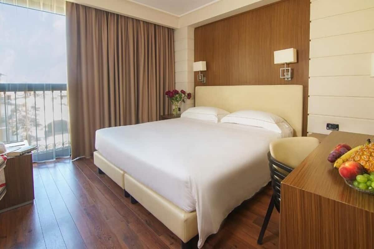 A hotel room with a large bed, a wooden desk with a fruit bowl, a chair, and a view of the outdoors.