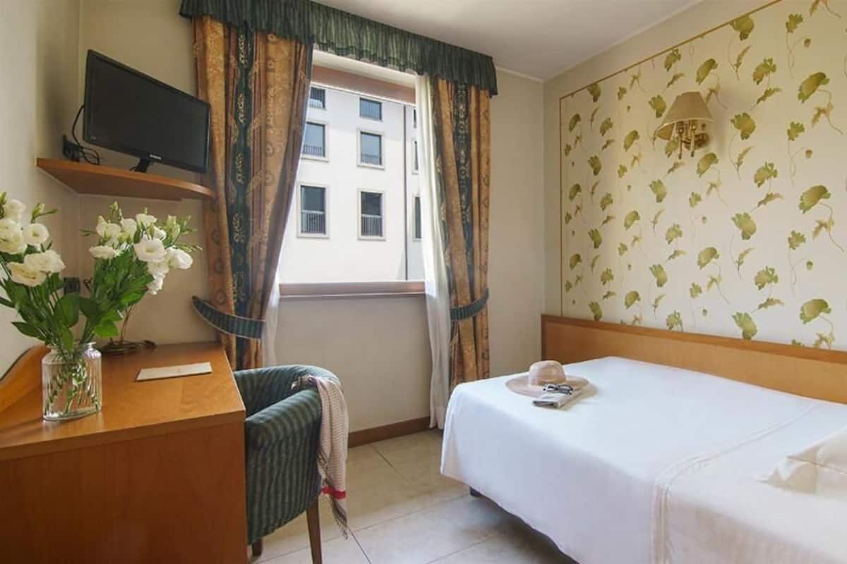 A hotel room with a bed, a desk with a television, a chair, a window with curtains, and a wall with a floral pattern.