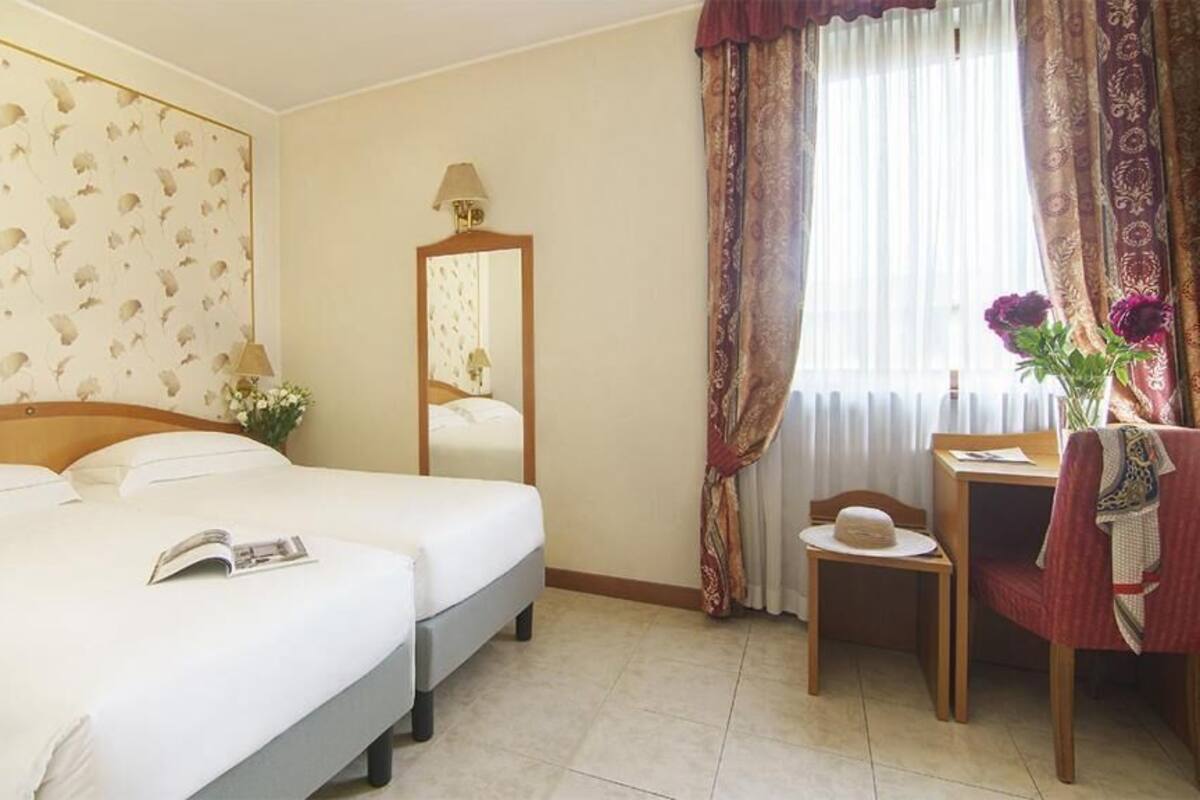 A hotel room with two beds, a desk, a chair, a mirror, and a window with curtains.