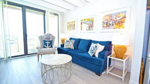 A living room with a blue sofa, round coffee table, and a chair with an anchor pillow. There are three framed pictures on the wall and a lamp on a side table. - Jada 2B-Oceanside 77th St, Free WiFi, Pool, Elev, W/D, AC (Ocean City)