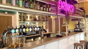 A bar area with a neon 'DRINKS!' sign, shelves of bottles, a tap bar, and wicker stools. - The People Caen (Caen)