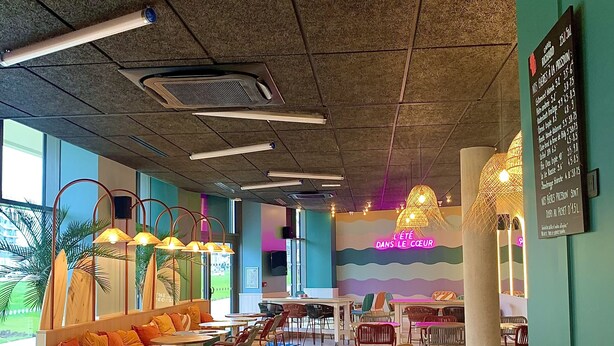 A spacious indoor area with tables and chairs, a high ceiling with lighting fixtures, and a colorful mural on the wall. - The People Caen (Caen)