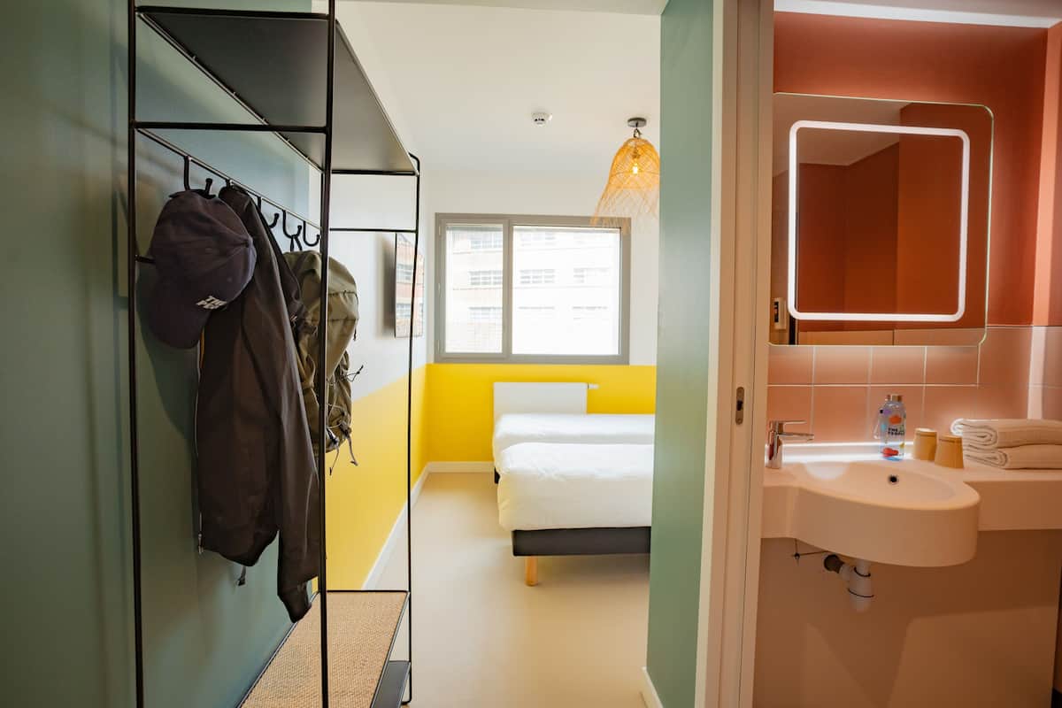 Ein Hotelzimmer mit Bett, Badezimmer mit Waschbecken und Spiegel, Kleiderständer mit Jacken und ein Fenster mit Blick auf Gebäude.