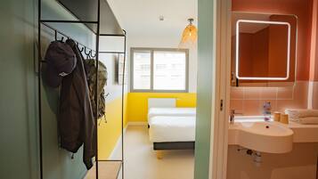 A hotel room with a bed, a bathroom with a sink and mirror, a coat rack with jackets, and a window with a view of buildings.