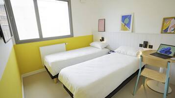 A small hotel room with two single beds, a round wooden table with a laptop, a window with blinds, and a wall with framed artwork.