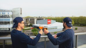 Two people toasting with drinks on a balcony overlooking a modern building with a red sign. - The People Caen (Caen)