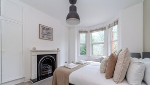 A bedroom with a fireplace, a bed with multiple pillows, a hanging light fixture, and a framed picture on the wall. - The Harlesden Villas (London)