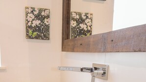 Een badkamer met een grote witte wastafel, een houten aanrechtblad, twee gestreepte vazen, een spiegel en bloemkunst aan de muur.