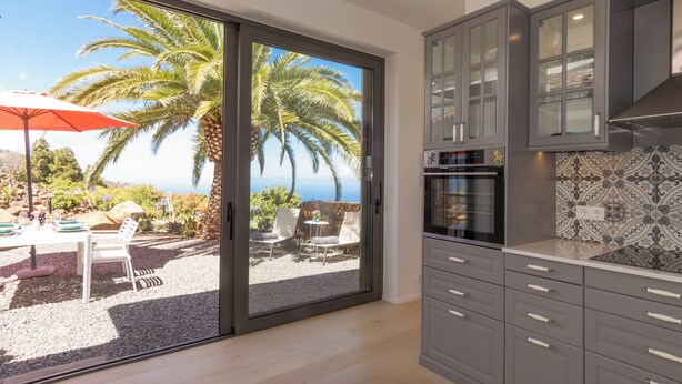 Een keuken met grijze kastjes, een betegelde spatwand en een schuifdeur naar een buitenzitgedeelte met een tafel, stoelen en een rode parasol.