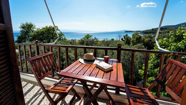 Een houten tafel met stoelen op een balkon met uitzicht op zee en heuvels.