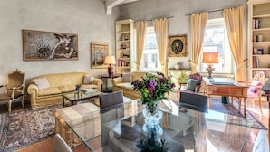 A spacious living room with a sofa, coffee table, and a glass dining table. There are bookshelves, a lamp, and a vase with flowers on the table.