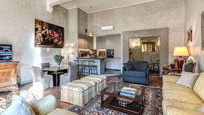 A spacious living area with a sofa, ottoman, coffee table, and a painting on the wall. A kitchen is visible in the background with a dining area.