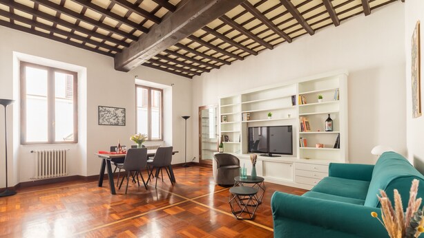 A spacious living area with a wooden floor, a teal sofa, a dining table with chairs, and a TV on a white shelf.