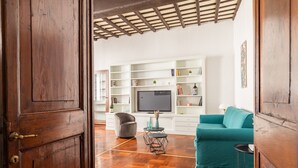 A living room with a wooden floor, a teal sofa, a grey armchair, a TV, and white shelves with various items.