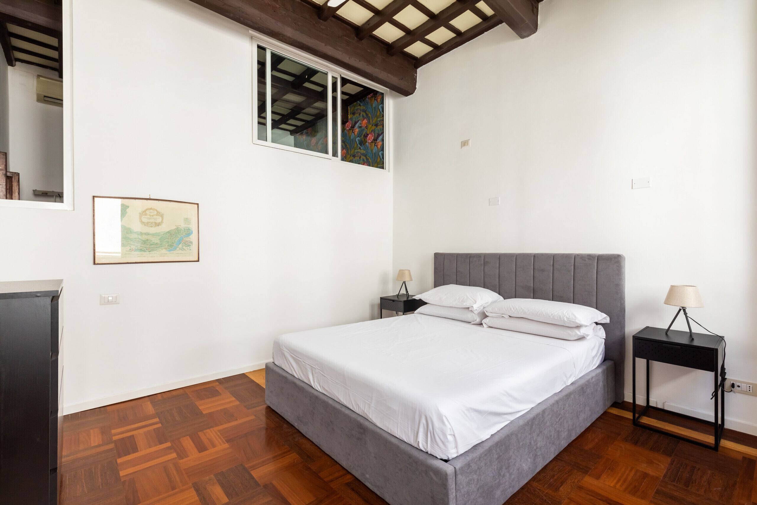 Ein Schlafzimmer mit einem Bett, einem Nachttisch, einem Fenster mit Blumenmuster und einem gerahmten Bild an der Wand.