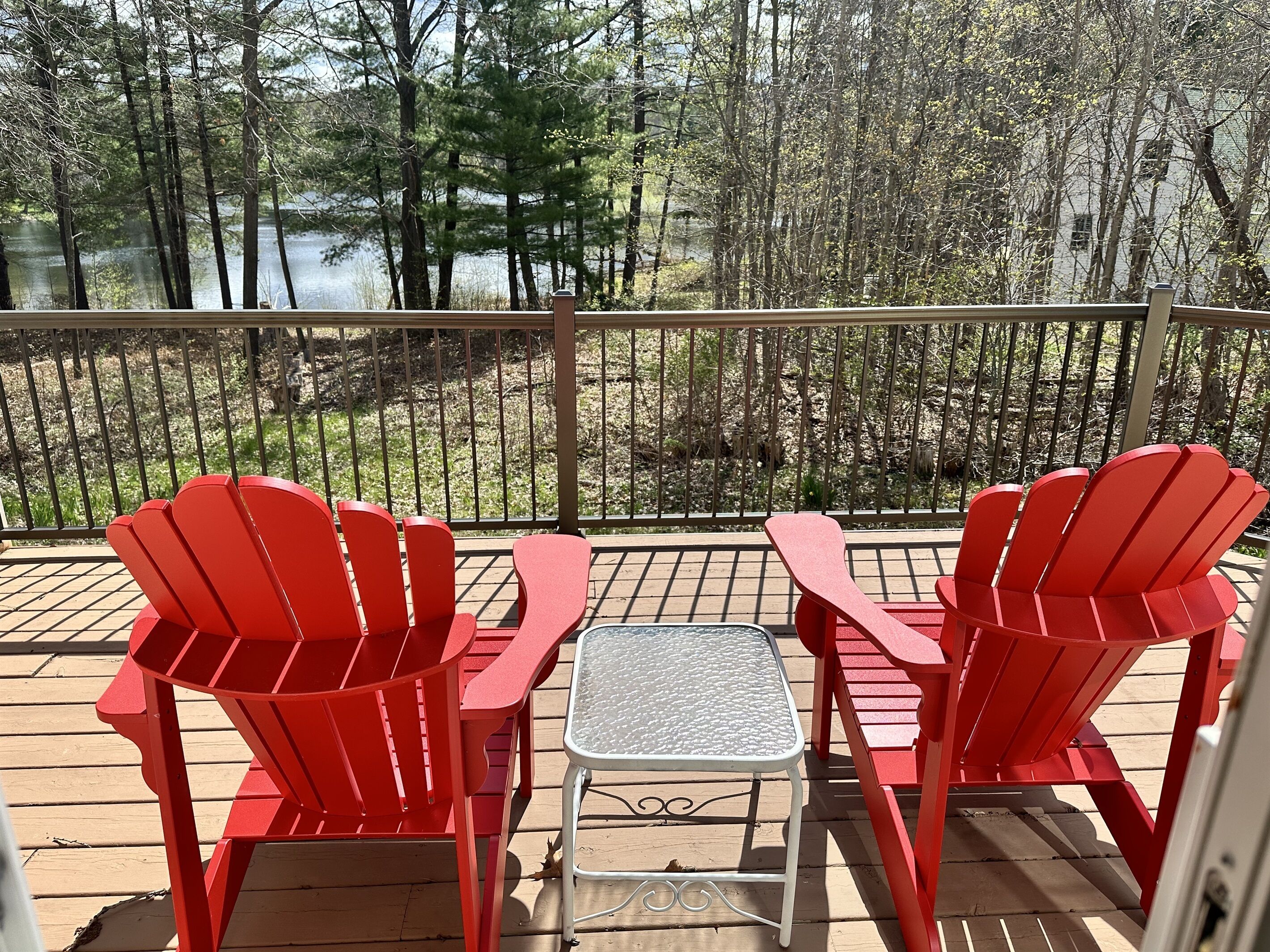 A deck with two red Adirondack chairs and a small table, overlooking a forested area and a body of water.