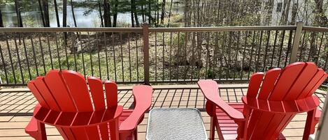 A deck with two red Adirondack chairs and a small table, overlooking a forested area and a body of water.