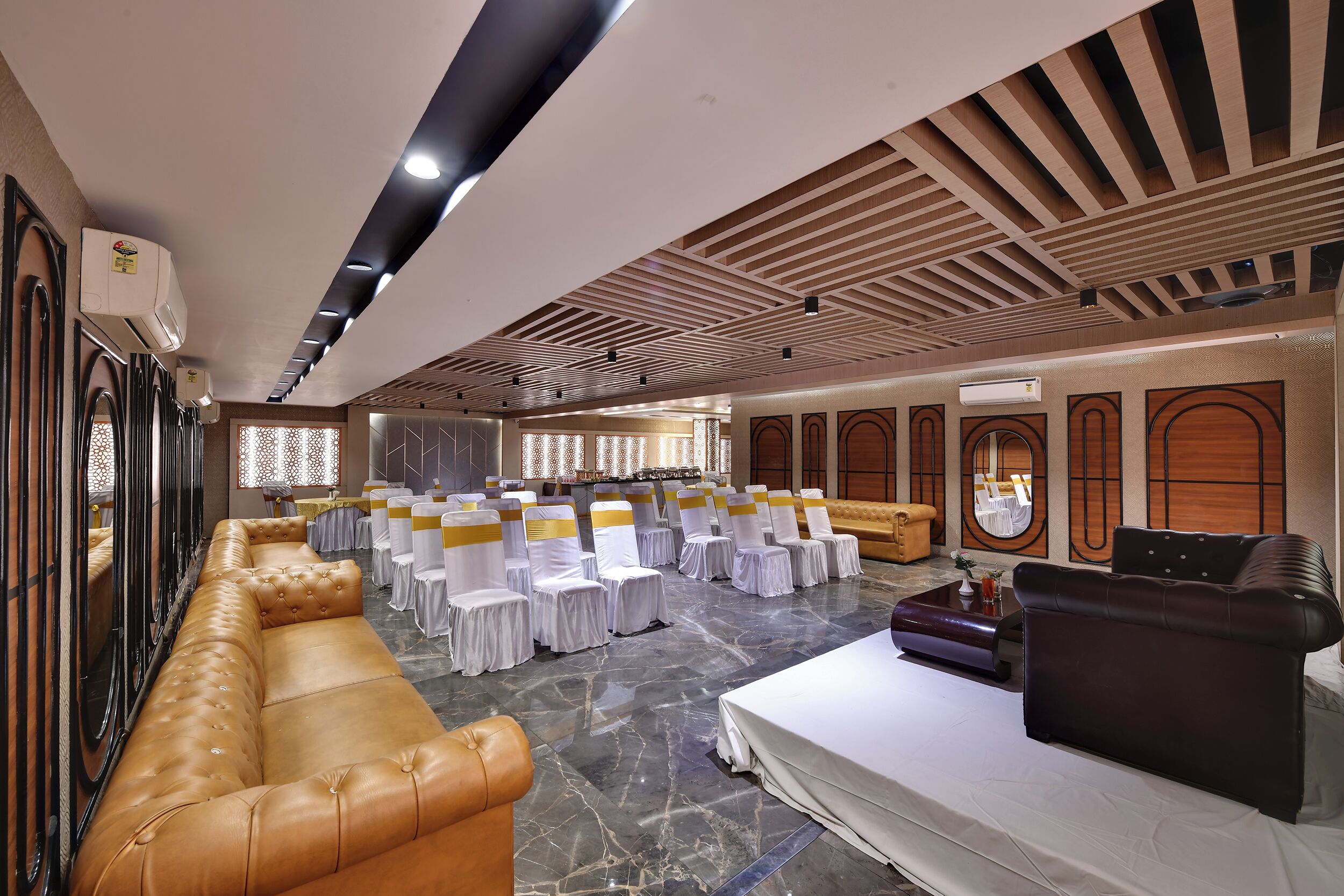 A spacious hall with rows of white-covered tables and chairs, a leather sofa, and a stage area.
