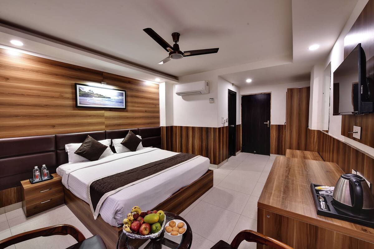 A modern hotel room with a wooden headboard, a bed with white linens, a flat-screen TV, a ceiling fan, and a breakfast bar with a kettle.