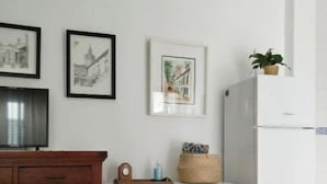 A kitchen with a wooden cabinet, a white table, and two white chairs. There are framed pictures on the wall and a plant on top of the refrigerator. - Apartamento Barrio Santa Cruz, Centro Sevilla (Sevilla)