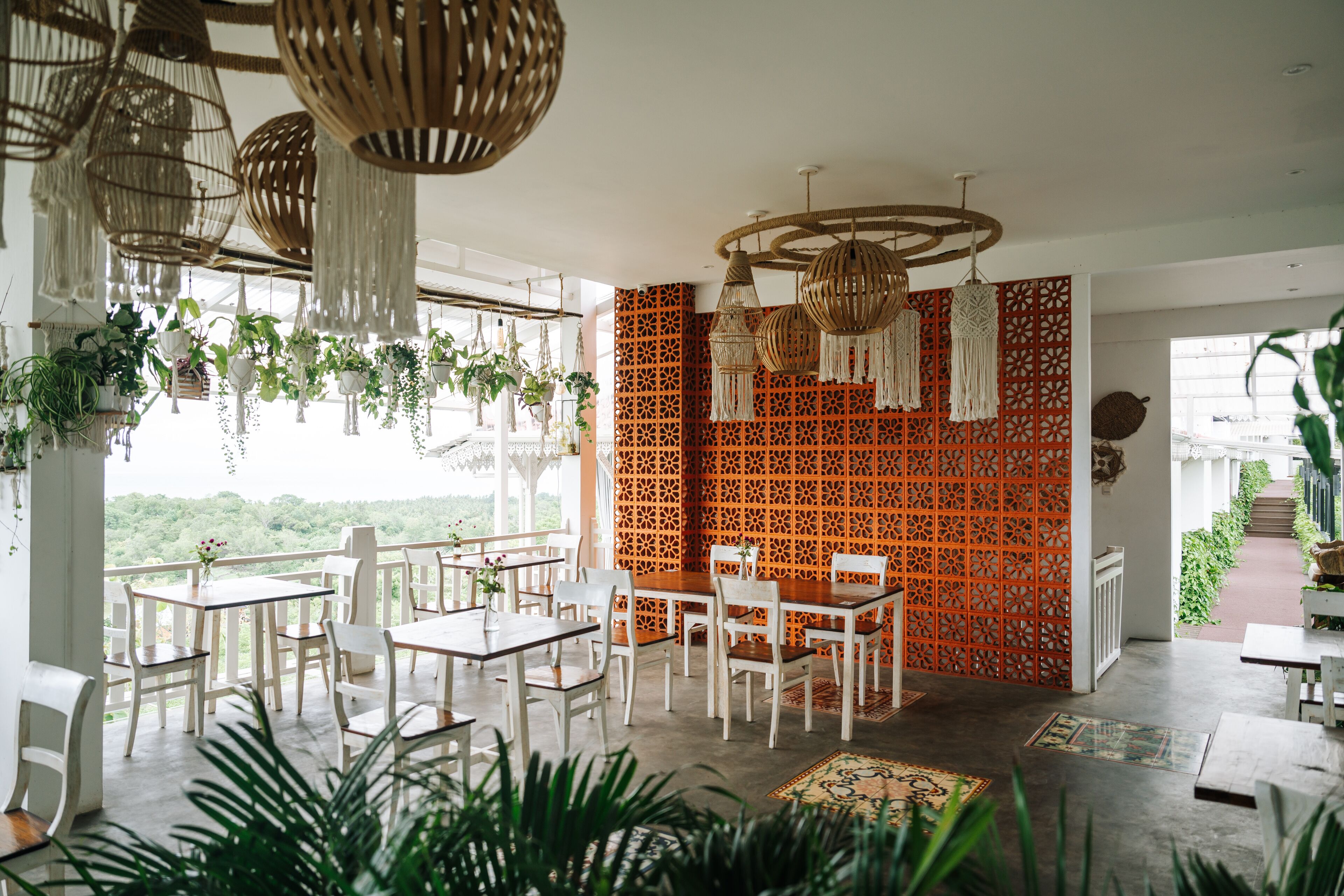 Un restaurante con mesas y sillas blancas, plantas colgantes y una pared con diseño rojo.