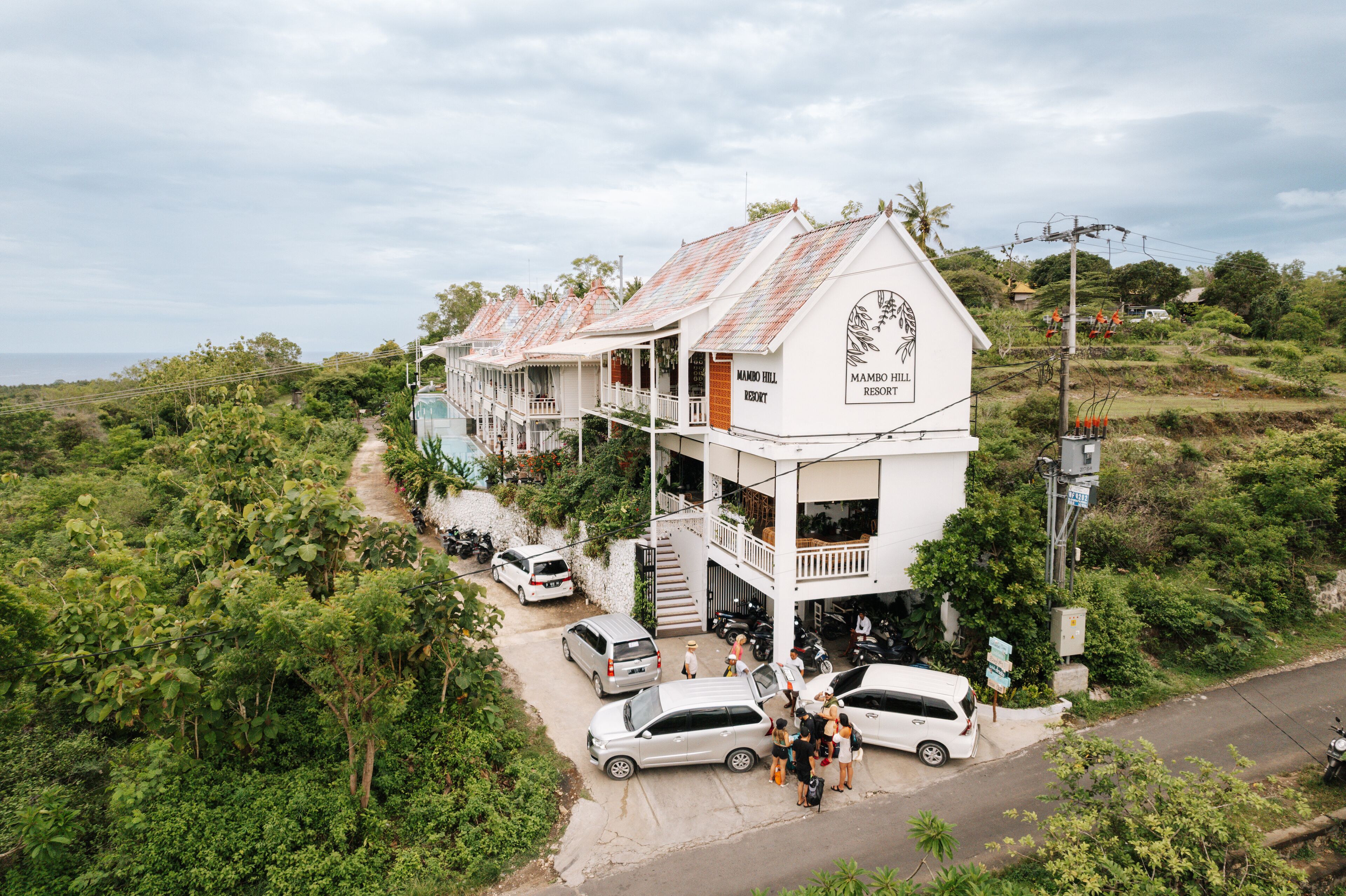 A row of white buildings with balconies, labeled "Mambo Hill Resort," surrounded by lush greenery.